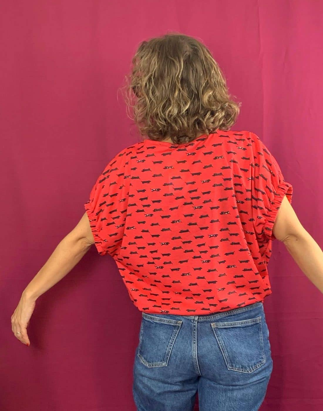 A person with wavy, shoulder-length hair faces away, arms slightly outstretched, wearing the La Compiano Lola T-Shirt in Sausage Print and blue jeans against a solid magenta background.