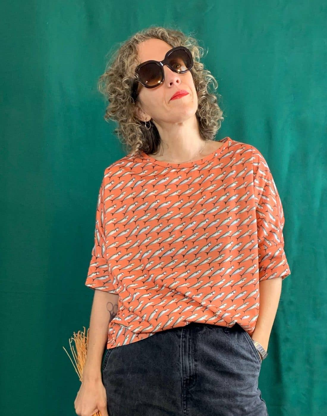 A woman with curly hair, wearing oversized sunglasses and La Compiano's vibrant orange Bond T-Shirt — Sardines Print with dark pants, stands before a green background, looking up in effortless Mediterranean style.