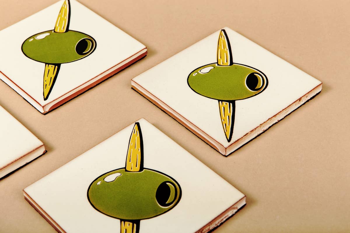 Four square ceramic Olivas Coasters by Bussoga feature a graphic green olive and yellow toothpick on a beige background. Arranged on a light brown surface, they add charm to your tabletop.
