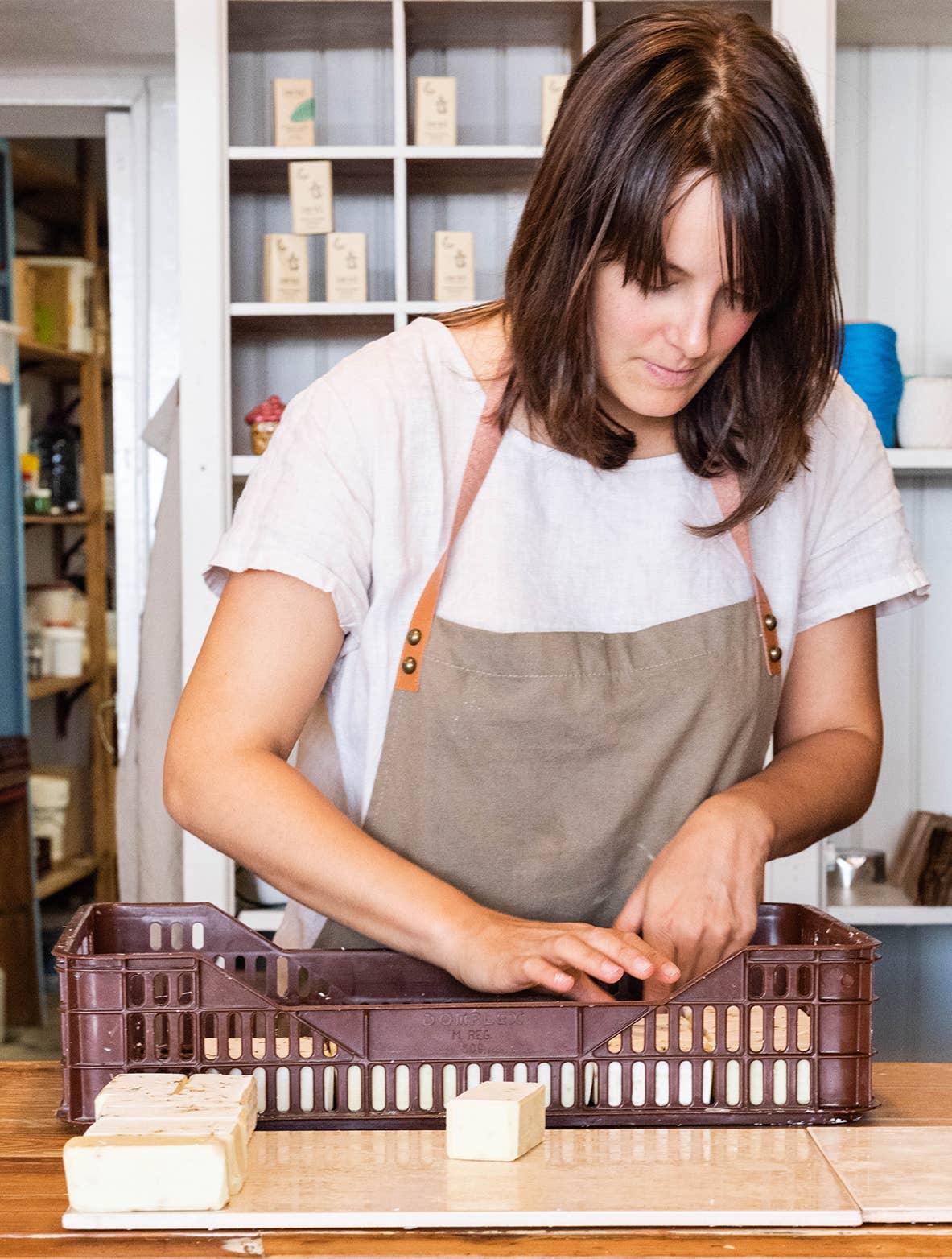 A woman in a light shirt and apron arranges Daki Olive Oil Soap with Ginger and Aloe Vera bars in a brown plastic crate on a wooden counter in a shop stocked with soaps and craft supplies.
