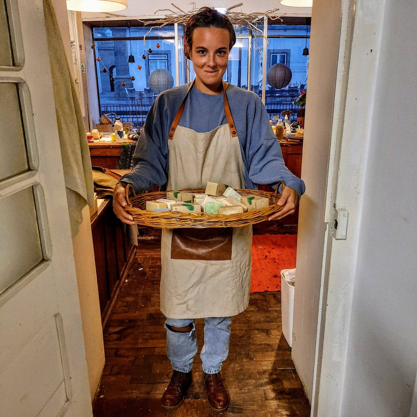 A person in an apron holds a large wicker basket filled with Daki Olive Oil Soap with Lavender. They stand indoors in a cozy room with wooden floors, a decorated window, and various items on display.