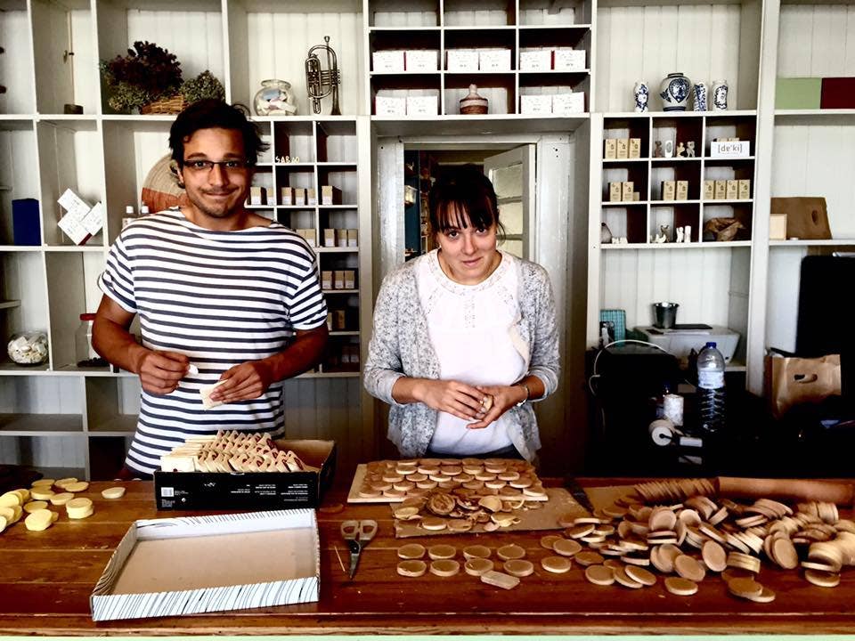 Two people stand behind a wooden counter stacking cookies and biscuits. Shelves behind them display items like Daki Olive Oil Soap with Roses and other natural products. Both face the camera amid a workspace full of baked goods.