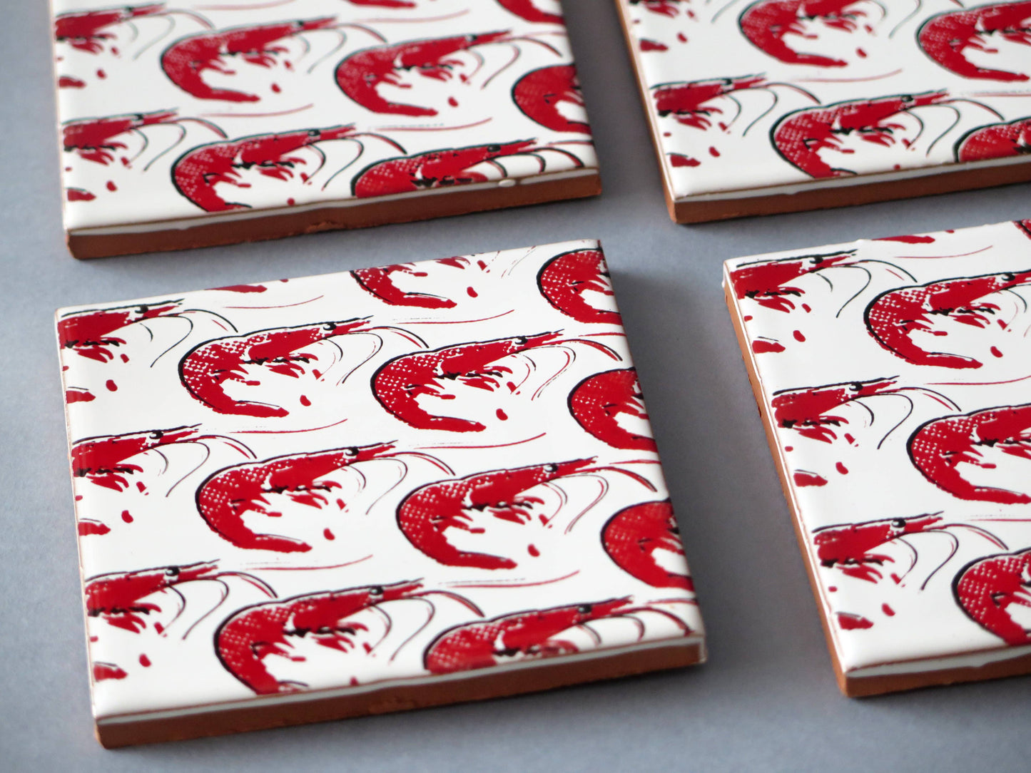 The Bussoga Gamba Design Ceramic Coasters set includes four handmade ceramic coasters from Spain, featuring a simple red shrimp pattern with scattered red splashes on a white background.