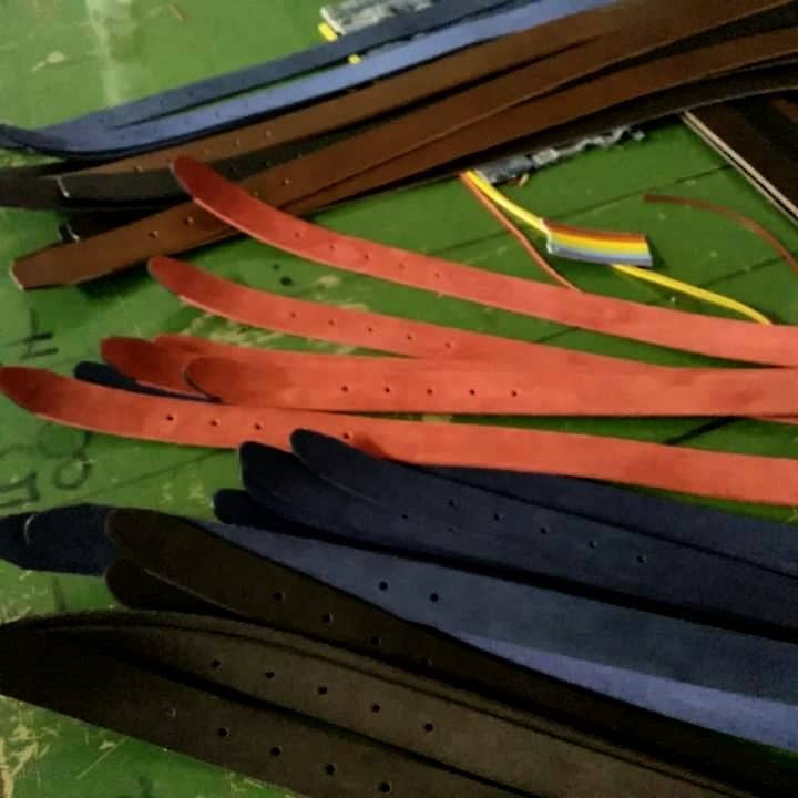 A close-up of several Creaciones Corpas Handcrafted Nubuck Leather Belt 3.5 cm in brown, red, and blue are laid out on a green surface, with tools in the background showcasing their Made in Spain craftsmanship.