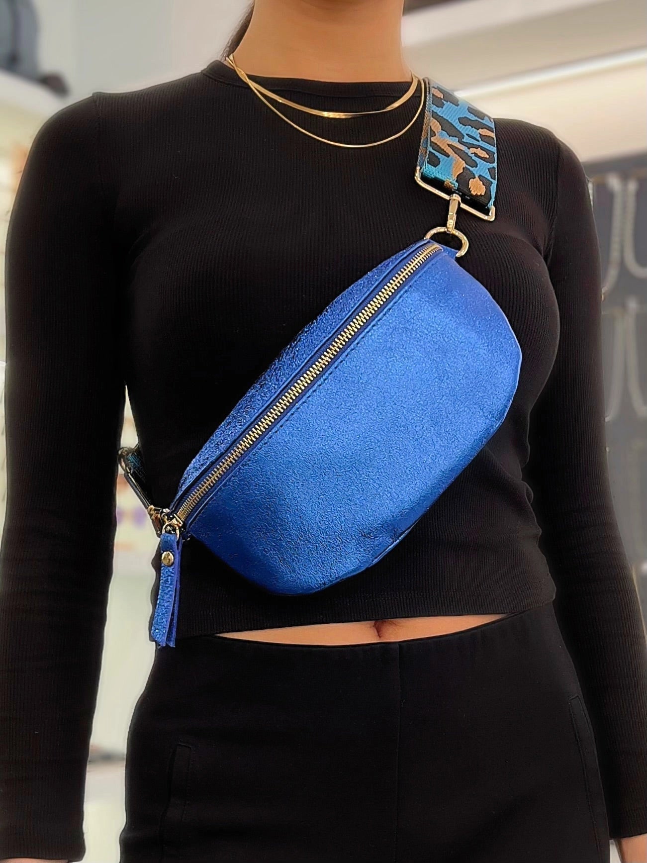 A woman in a black long-sleeve top and gold necklaces carries the Cassette Barcelona Leather Fanny Pack Crossbody Bag with a blue camouflage strap across her chest.