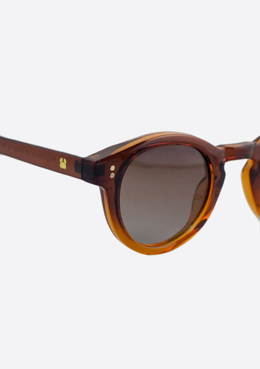 Close-up of Oceanides Sunglasses (Polarized) - Rodea Crystal Gradient Brown, featuring round frames, gradient brown polarized lenses, and a subtle gold logo on the arm, set against a plain white background.