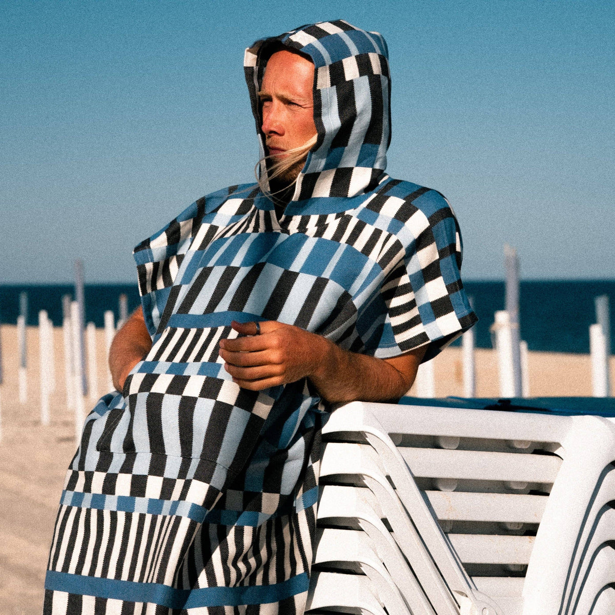 A person wearing the Guincho Poncho by Futah, featuring a blue, black, and white checkered cotton design, leans on stacked white lounge chairs at a sunny beach with sand and the ocean in the background.