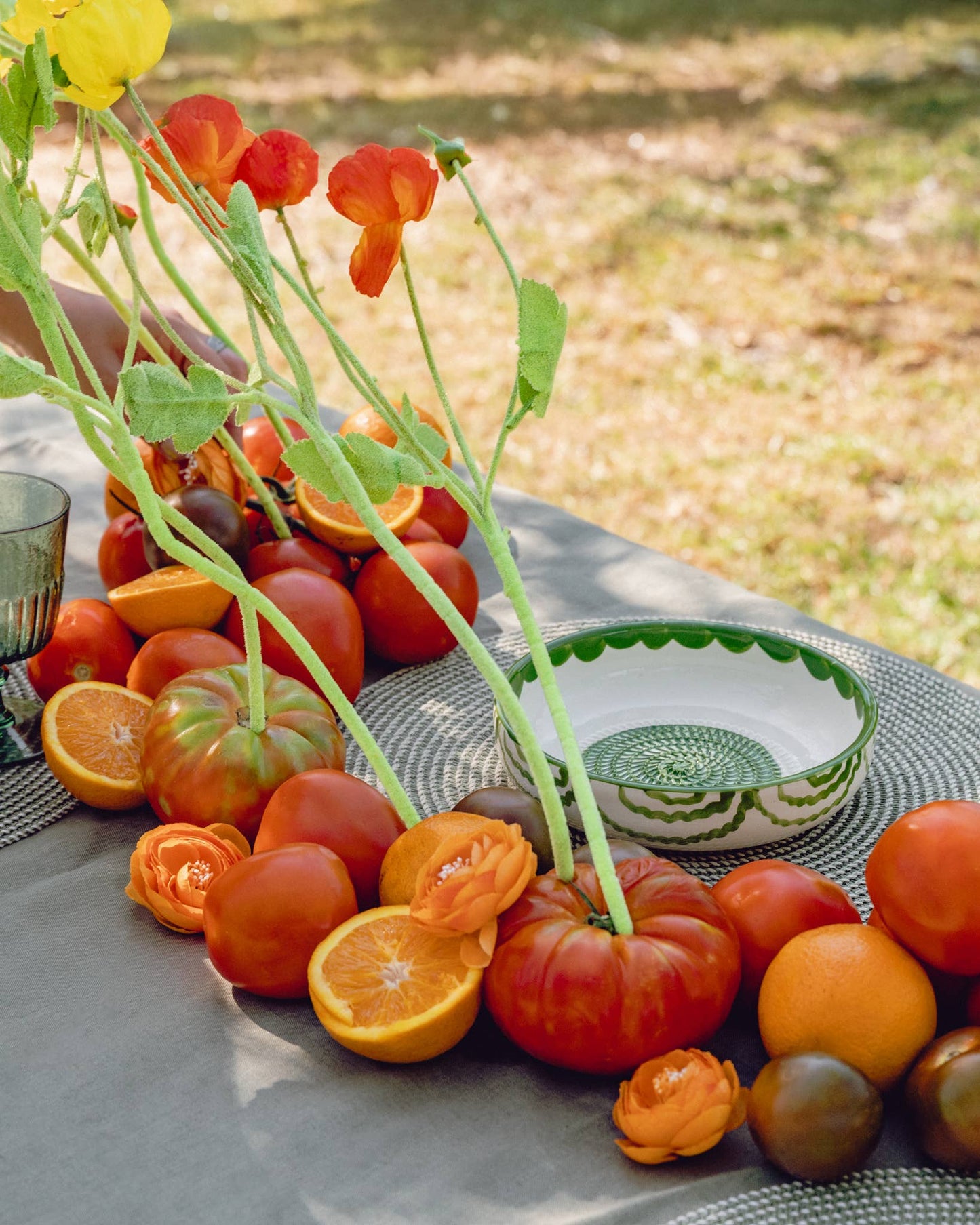 A table outdoors features tomatoes, sliced oranges, orange flowers, and upright green and yellow flowers next to the Iberica - Pretty things from Portugal Large Pottery Grater Plate - Roots, displayed on a gray tablecloth.