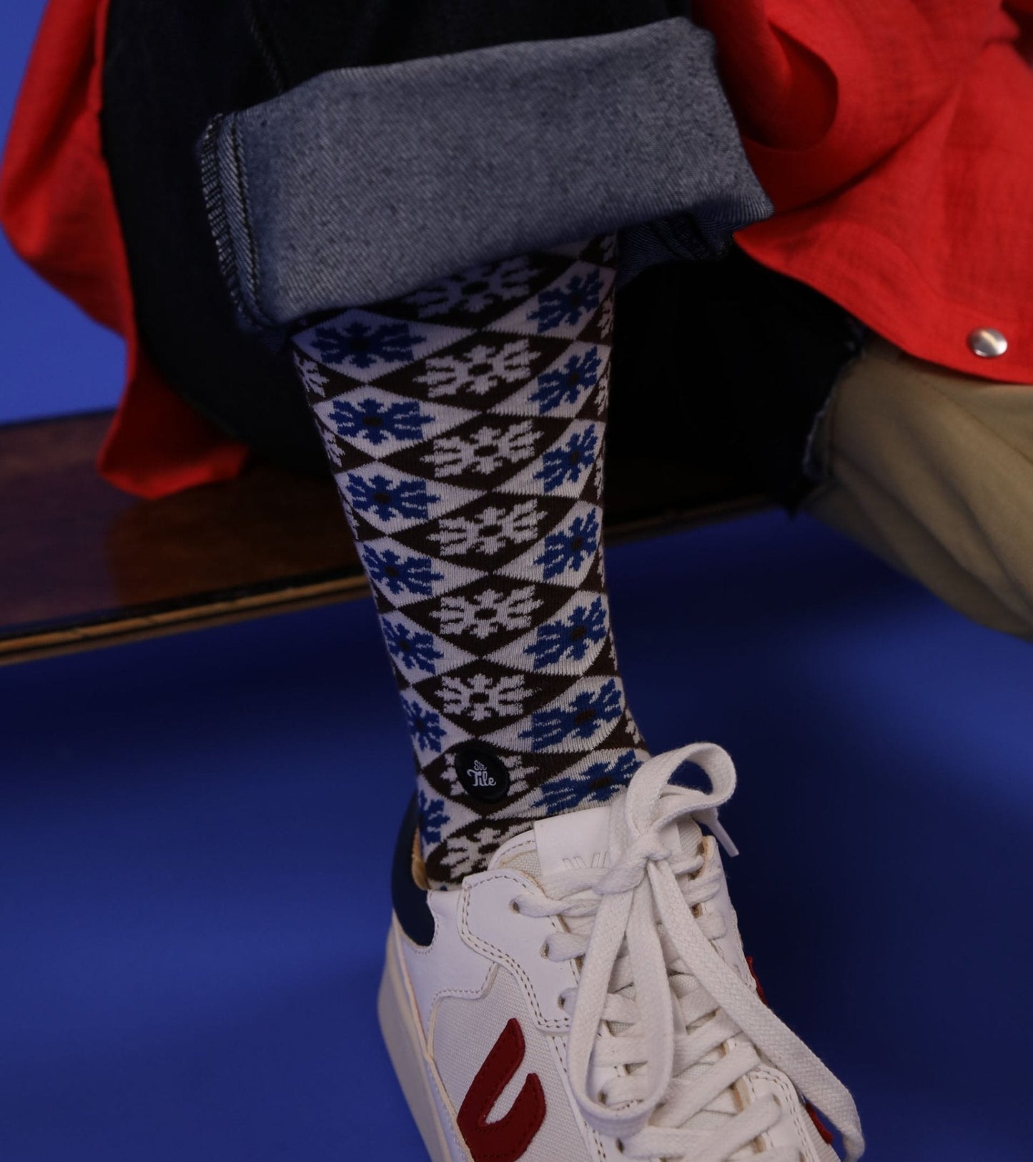 A person sits with one leg crossed, wearing unisex SirTile Stylized Floral socks made from Egyptian cotton featuring a bold geometric design. Beige pants and white sneakers complete the look, set against a vibrant blue background.