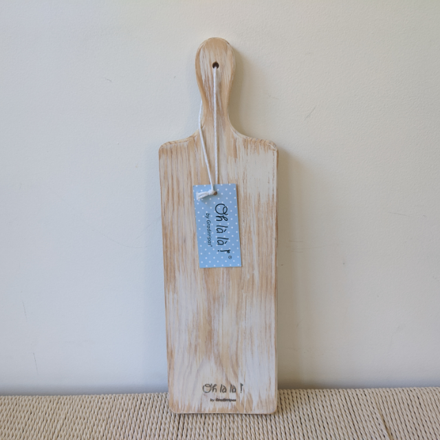 The Gradirripas Cheese Serving Board (60 x 22cm) is a handmade, light wooden pine board with a handle and blue polka-dotted “Shabat” tag, hanging by a string and standing upright on a woven surface against a light wall.