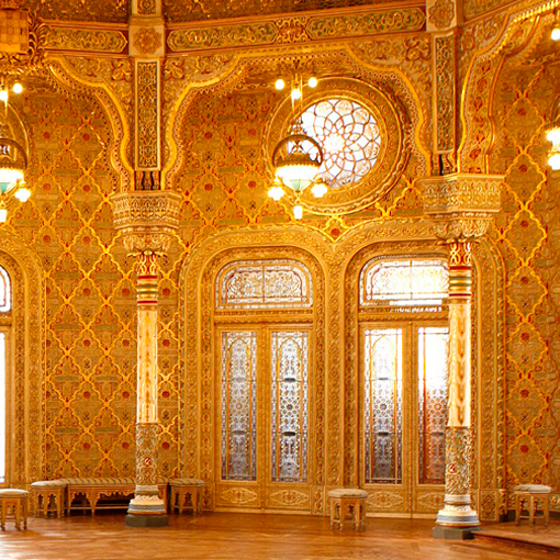 An ornate golden hall with Moroccan-style columns, patterned walls, and large arched windows glows under elegant chandeliers. A Terra Lusa Stained Glass Eco Shopper adds a touch of sustainable style to the scene.