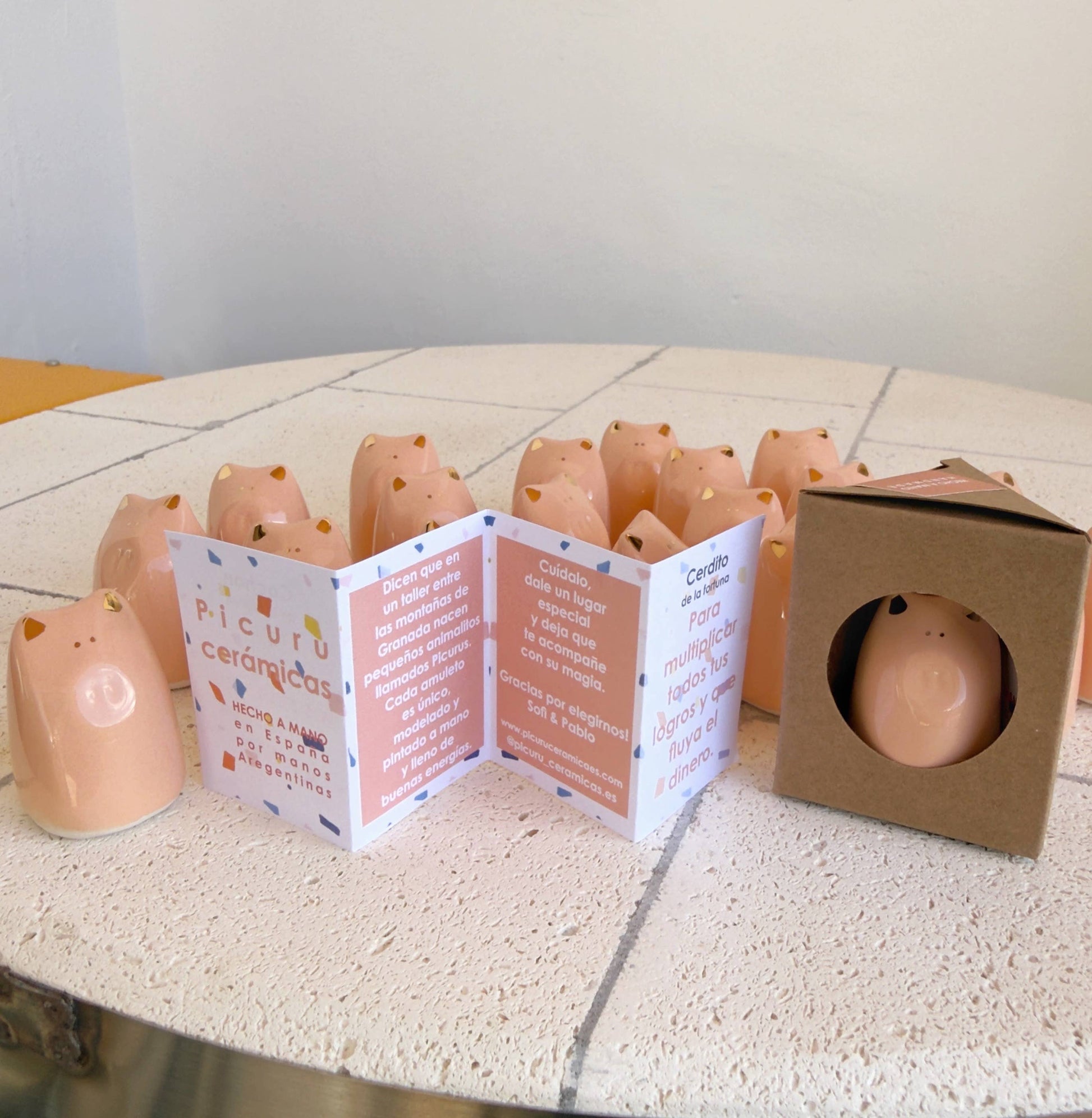 A display of Picuru ceramicas’ Fortune Pig Amulet - Gold Luster 5 Languages shows small pink piggy figurines on white tiles, with folded info cards and a boxed amulet featuring a cutout window in front.