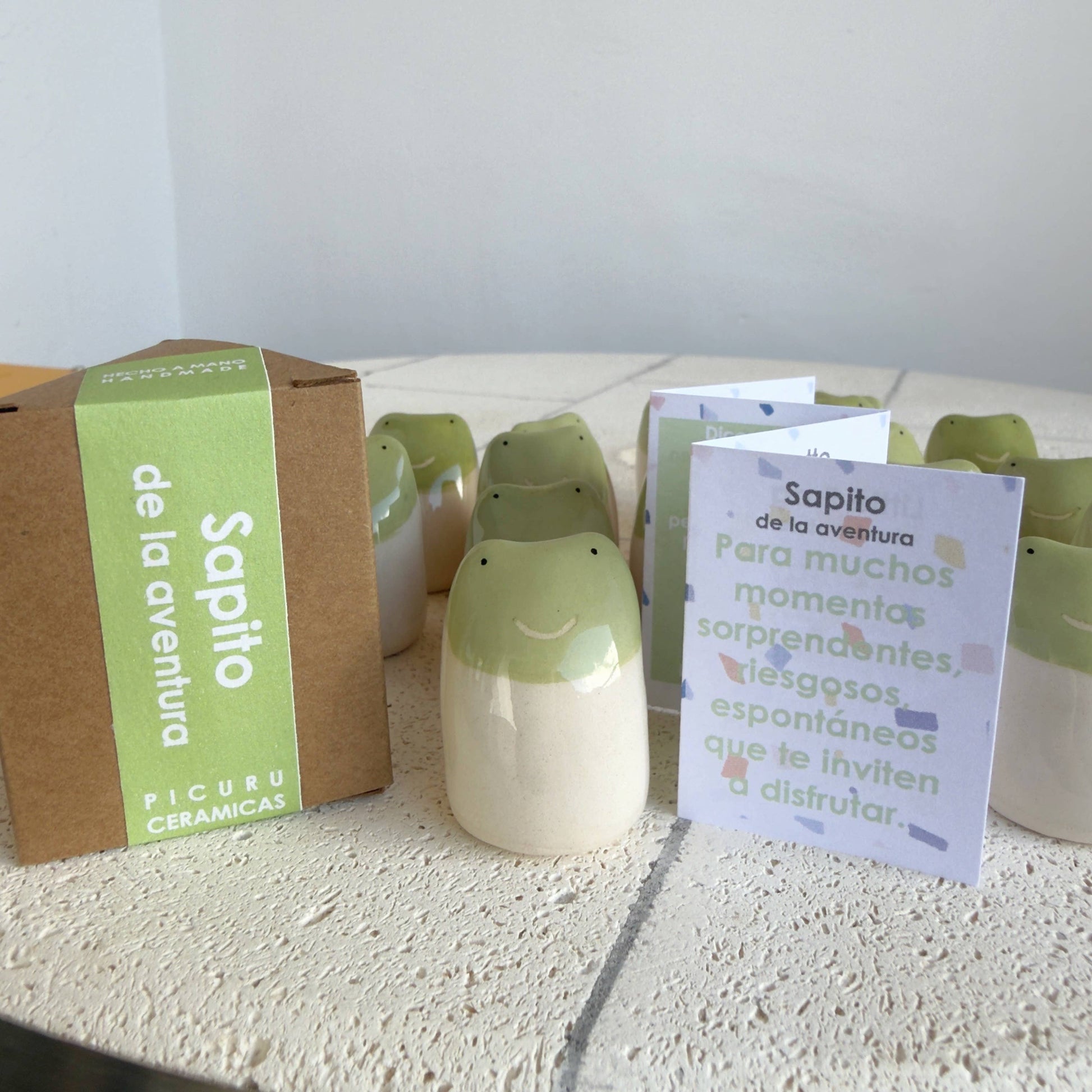 A set of Little Frog Amulet of Adventures by Picuru ceramicas, handmade with green-topped smiling faces, is displayed beside its brown box—making a unique gift and charming symbol of joy and new experiences.