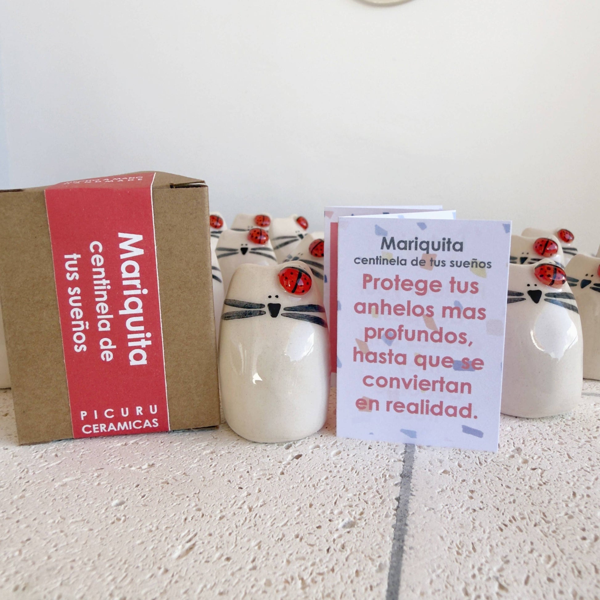 A set of Picuru ceramicas "Ladybug Amulet, Guardian of Your Dreams" ceramic cat figurines with ladybugs on their heads is displayed on a white surface. This handmade lucky charm comes with a Spanish message card, perfect as a good luck gift.