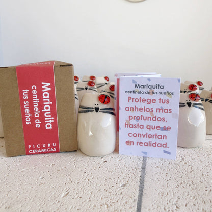 A set of Picuru ceramicas "Ladybug Amulet, Guardian of Your Dreams" ceramic cat figurines with ladybugs on their heads is displayed on a white surface. This handmade lucky charm comes with a Spanish message card, perfect as a good luck gift.