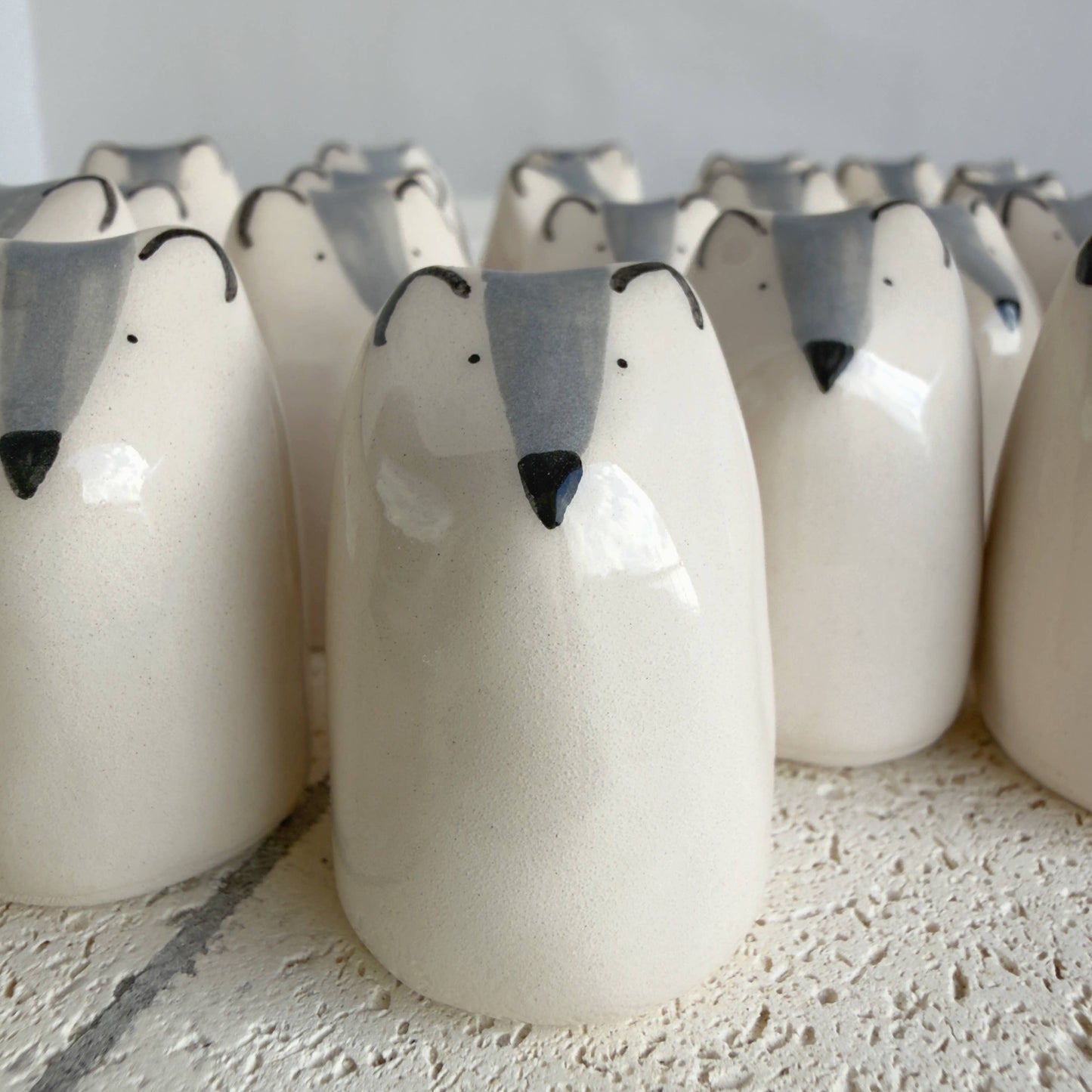 Rows of Picuru ceramicas' Little Wolf Amulet of Courage figurines—handmade symbols of strength and growth with white bodies, gray snouts, black noses, and simple dot eyes—are closely arranged on a textured surface.