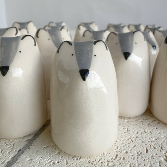 Rows of Picuru ceramicas' Little Wolf Amulet of Courage figurines—handmade symbols of strength and growth with white bodies, gray snouts, black noses, and simple dot eyes—are closely arranged on a textured surface.