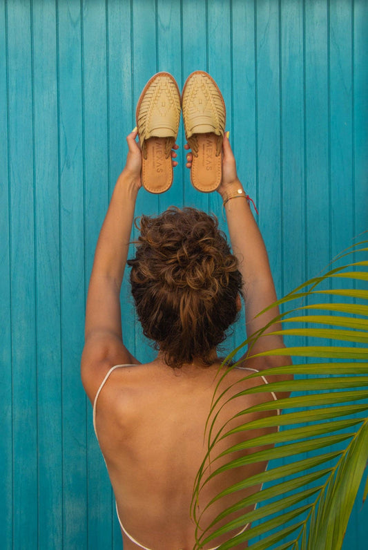 A person with curly hair and tan skin stands by a bright blue wooden wall, holding up Yellow San Pancho Slide Mules from Mexas EU. They wear a white strap top, with a green palm leaf in the foreground on the right.