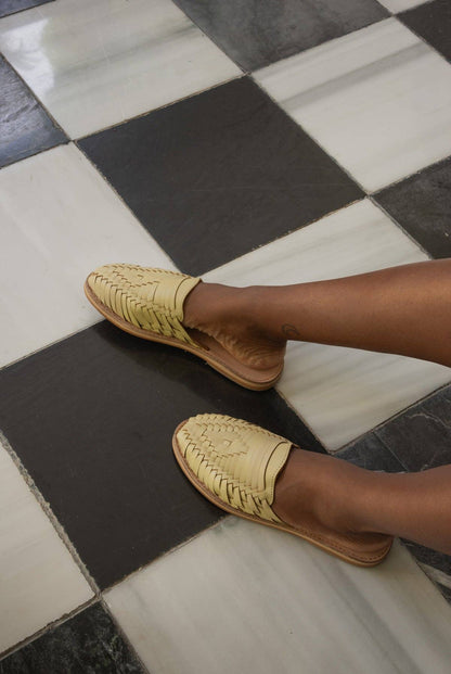 A person wears Mexas EU Yellow San Pancho Slide Mules with a cushioned footbed, resting their legs on a black-and-white checkered tile floor. Only the lower legs and feet are visible.