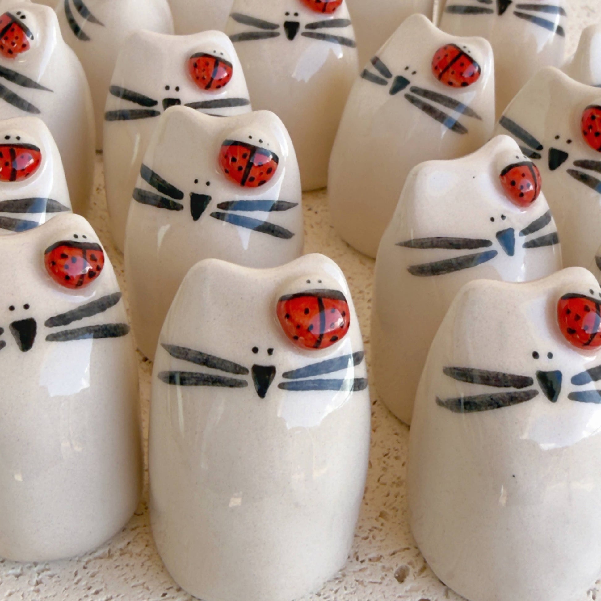 Picuru ceramicas’ "Ladybug Amulet, Guardian of Your Dreams" features handmade ceramic white cat figurines with black whiskers and a red ladybug charm painted on their heads, arranged closely together on a textured surface.