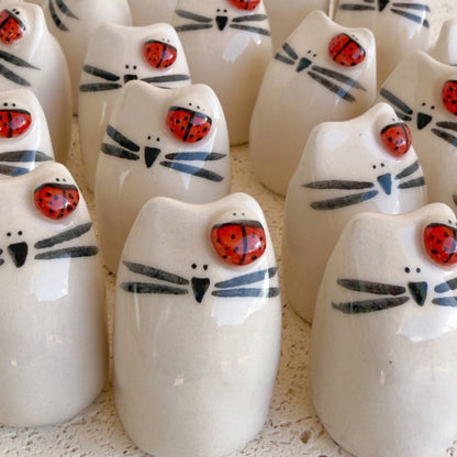 Picuru ceramicas’ "Ladybug Amulet, Guardian of Your Dreams" features handmade ceramic white cat figurines with black whiskers and a red ladybug charm painted on their heads, arranged closely together on a textured surface.