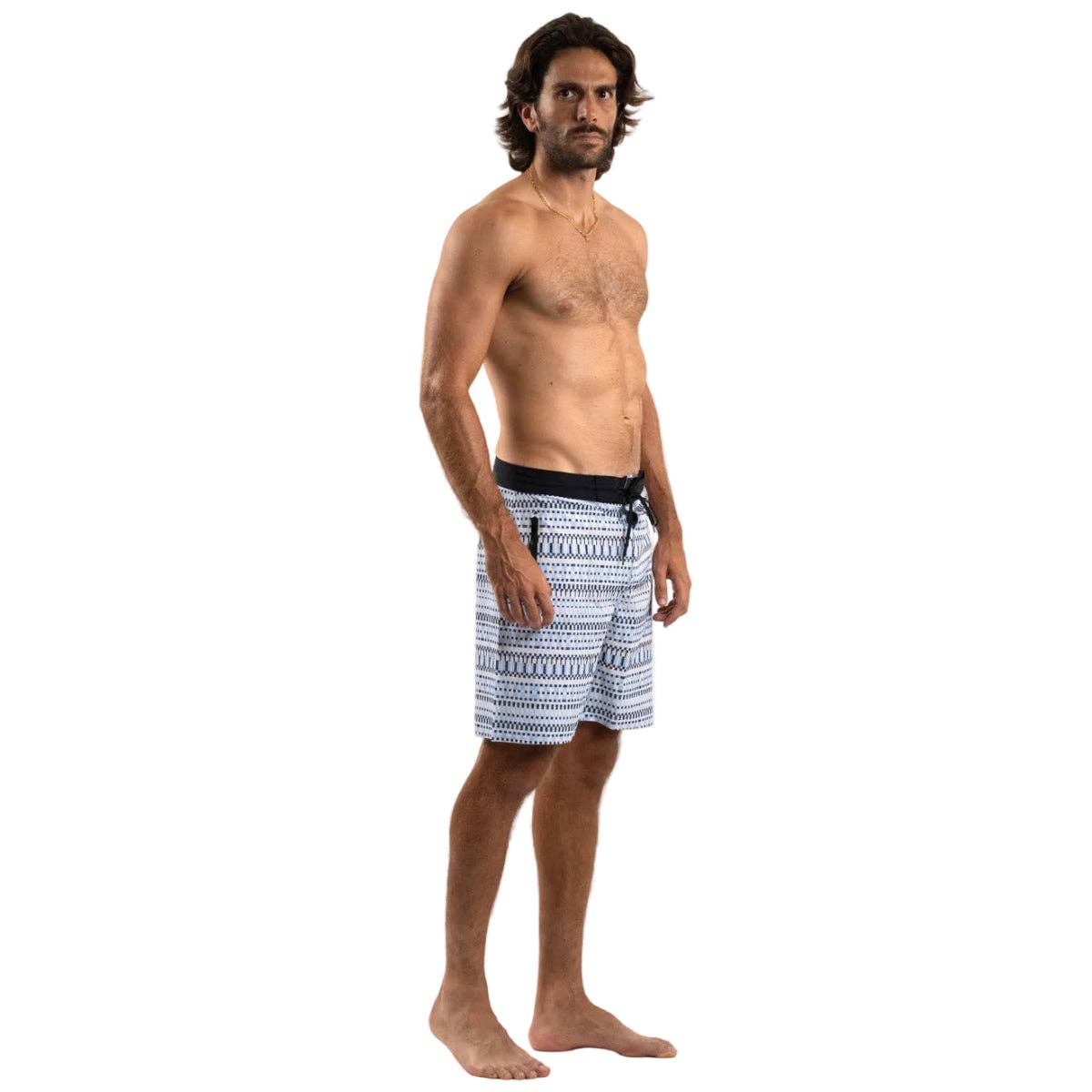 A shirtless man with medium-length brown hair stands barefoot against a plain white background, wearing Futah’s Cacela Blue Boardshorts featuring a drawstring waist and patterned design.