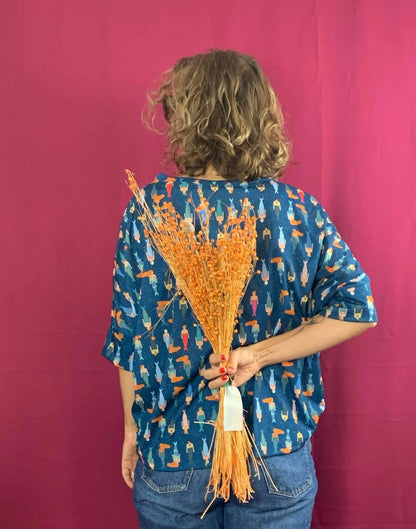 A person with curly hair faces away, wearing La Compiano’s Bond T-Shirt - Mermaid Print, featuring a vibrant pattern. They hold dried orange flowers behind their back against a solid magenta background, evoking Mediterranean style.