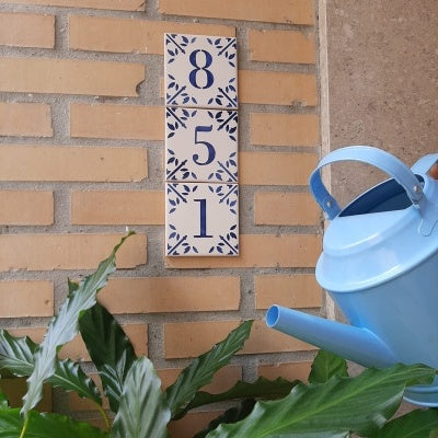 A blue watering can is beside a green plant in front of a brick wall, decorated with TilePassion's Personalized Portuguese Tile House Number sign—handpainted tiles displaying the numbers 8, 5, and 1 arranged vertically.