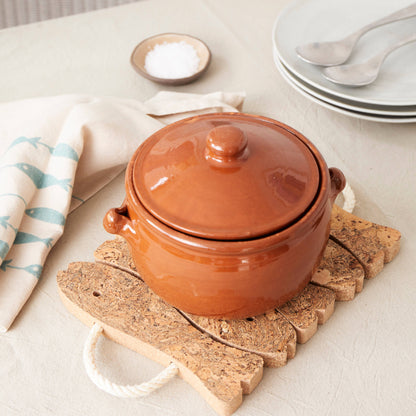 A closed brown ceramic pot sits on a LIGA Natural Cork Trivet - Fish, next to a folded napkin, a small dish of salt, and stacked plates with spoons, all atop a light-colored tablecloth.