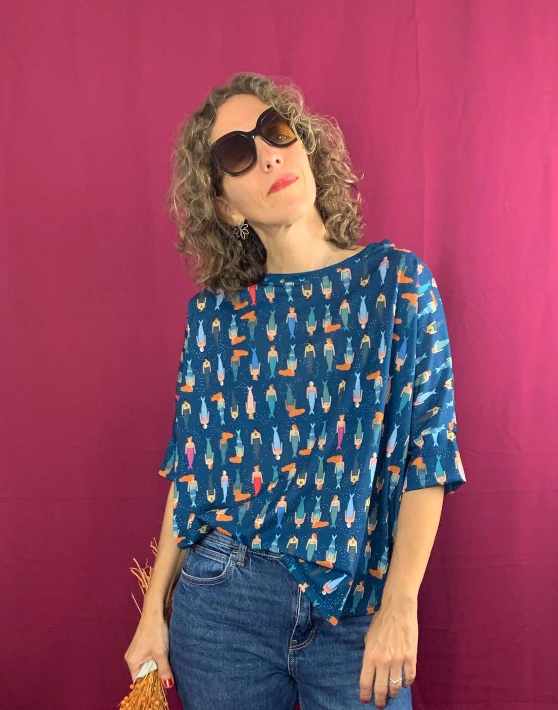 A woman with curly hair, large sunglasses, and a La Compiano Bond T-Shirt - Mermaid Print, stands before a magenta background, holding a woven handbag and gazing upward and to the side.