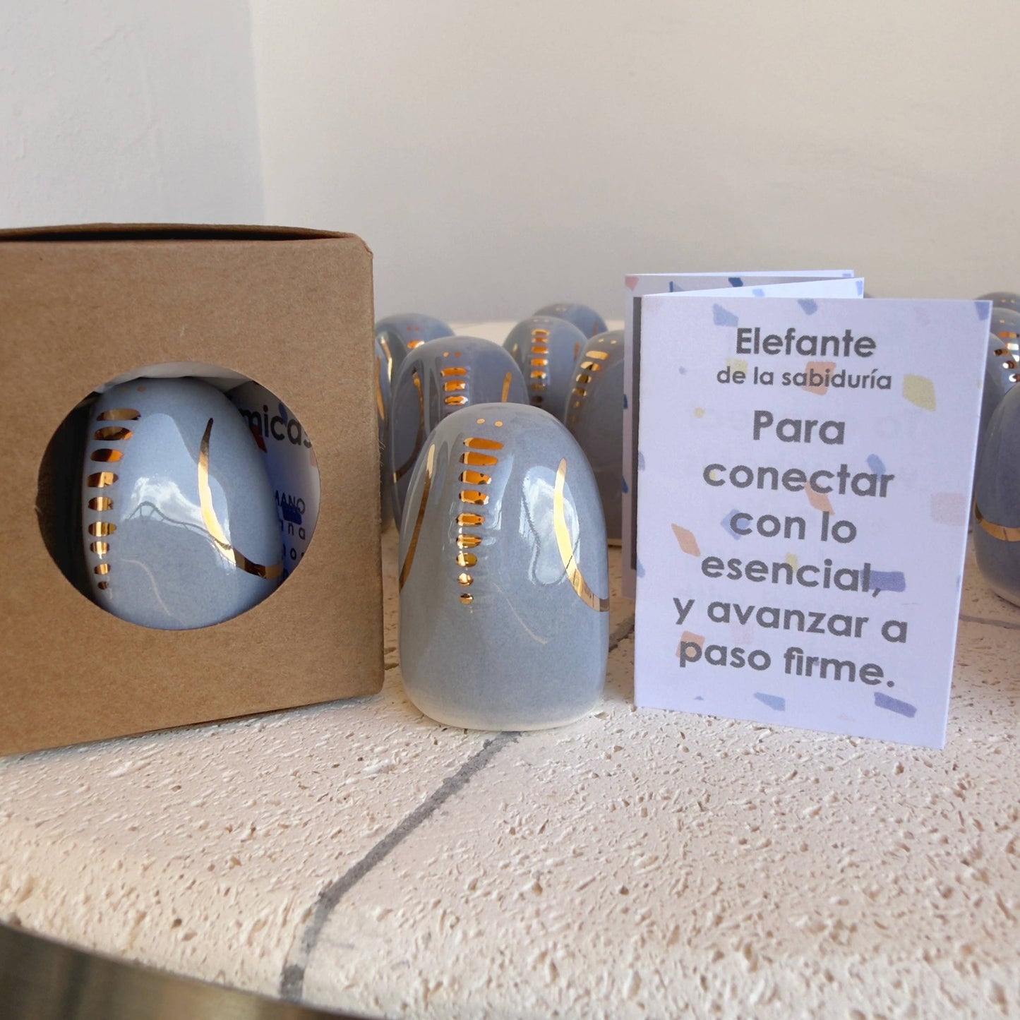 The Amule Elephant of Wisdom "Gold" by Picuru ceramicas is a handmade gray ceramic figurine with gold accents, shown beside a Spanish card. Several more appear in the background. Available in 5 languages, it makes a meaningful gift.