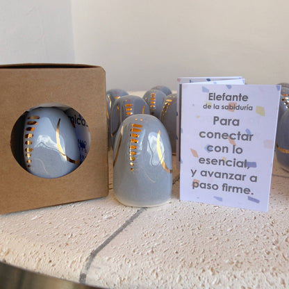 The Amule Elephant of Wisdom "Gold" by Picuru ceramicas is a handmade gray ceramic figurine with gold accents, shown beside a Spanish card. Several more appear in the background. Available in 5 languages, it makes a meaningful gift.