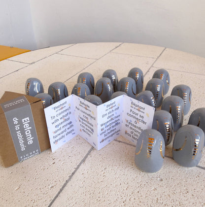 A group of Picuru ceramicas Amule Elephant of Wisdom "Gold" figurines sits on a light surface beside folded cards with text in 5 languages and cardboard packaging labeled Elefant de la solidaritat.