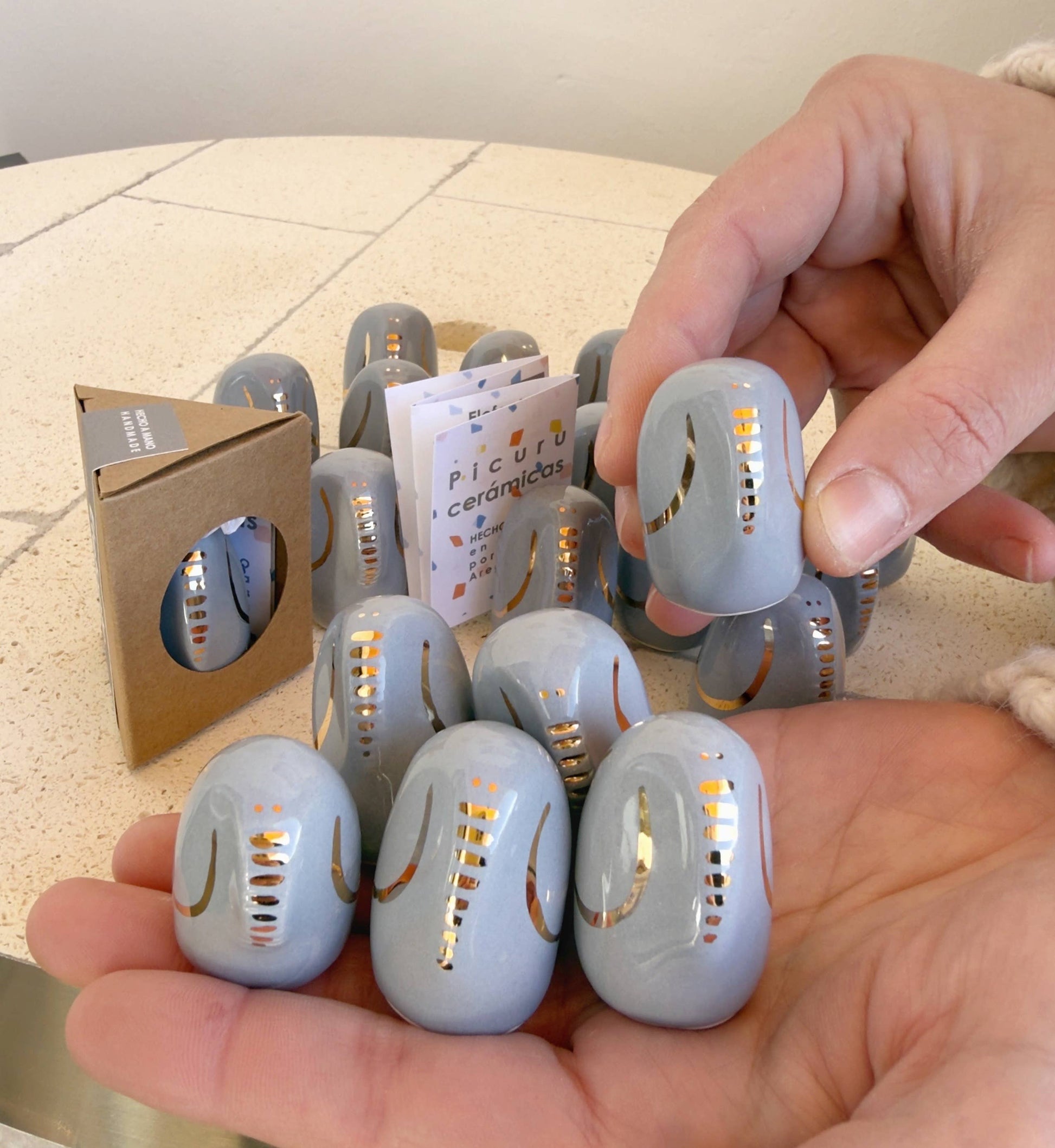 A person holds several light-gray Amule Elephant of Wisdom "Gold" talisman stones by Picuru ceramicas, with shiny gold accents. More stones and a brown box with a booklet are on the table, making this a thoughtful gift in 5 languages.