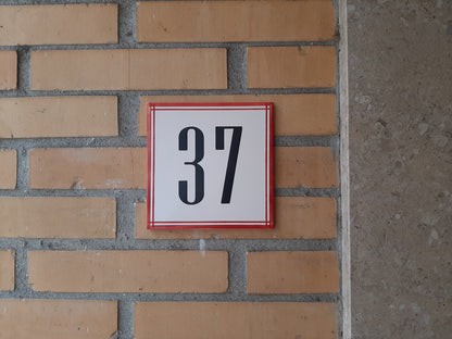 A white square TilePassion Handpainted Ceramic Door Number (37) in bold black font with a red border, styled like custom Portuguese tiles, is mounted on a yellow brick wall beside gray stone.