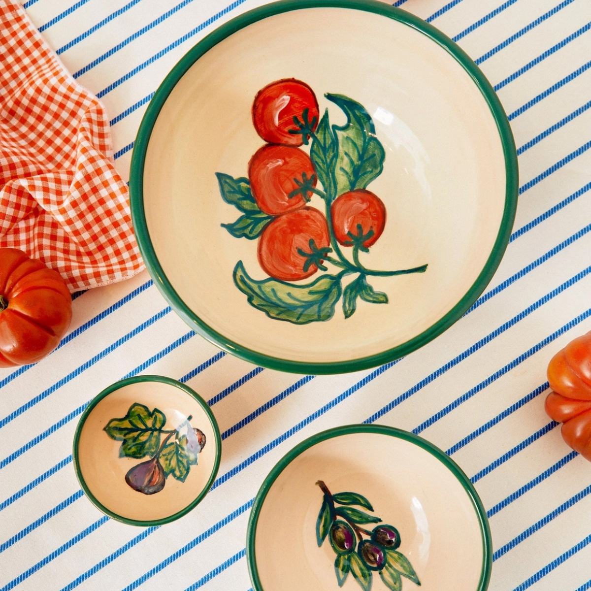 Three Eturel Madrid Huerta Stamped Ceramic Bowls, hand-painted with tomatoes, figs, and olives, are displayed on a blue-striped tablecloth with an orange-checkered napkin and fresh tomatoes, highlighting their rustic charm for any setting.