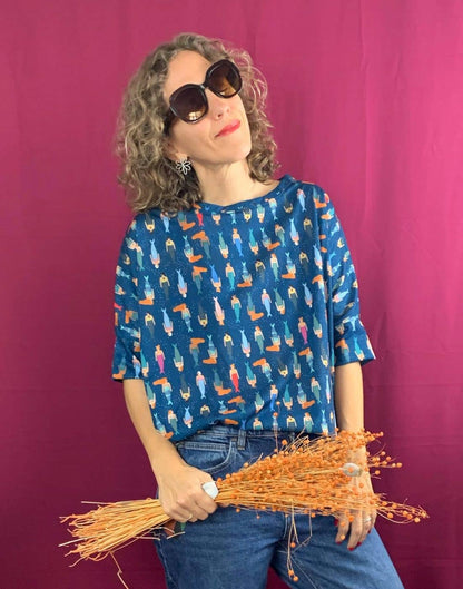 A woman with curly hair, wearing large sunglasses and the La Compiano Bond T-Shirt - Mermaid Print with jeans, stands against a magenta background, holding dried orange flowers and smiling softly.