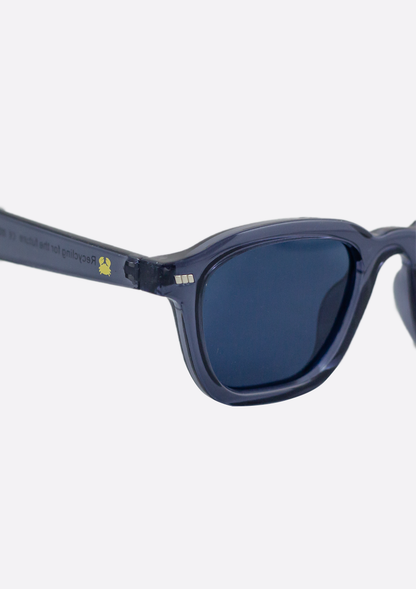 Close-up of Oceanides Neda Crystal Blue Sunglasses featuring rectangular polarized lenses, a small yellow turtle icon on the recycled plastic frame, and partially visible text.