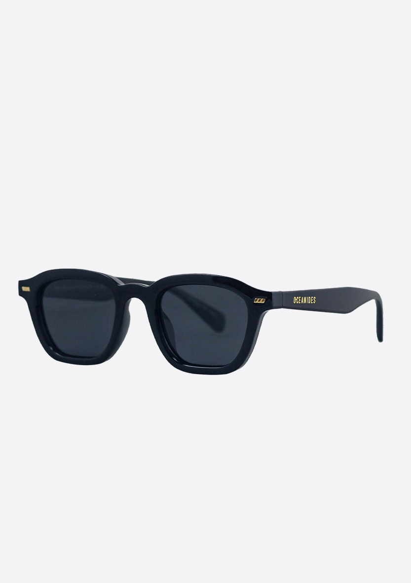 The Oceanides Neda Shiny Black Sunglasses feature a thick recycled plastic frame, dark polarized lenses, and gold DETAINED accents on the arms, all displayed against a plain white background.