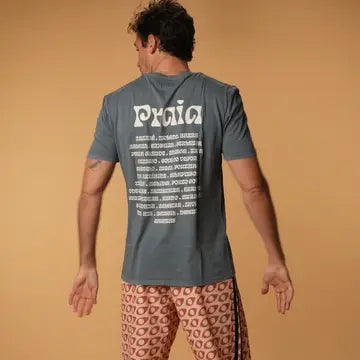 A man, seen from the back, wears the Futah Praia T-Shirt with beach-themed text on the back and pairs it with pink patterned shorts, set against a plain beige background.