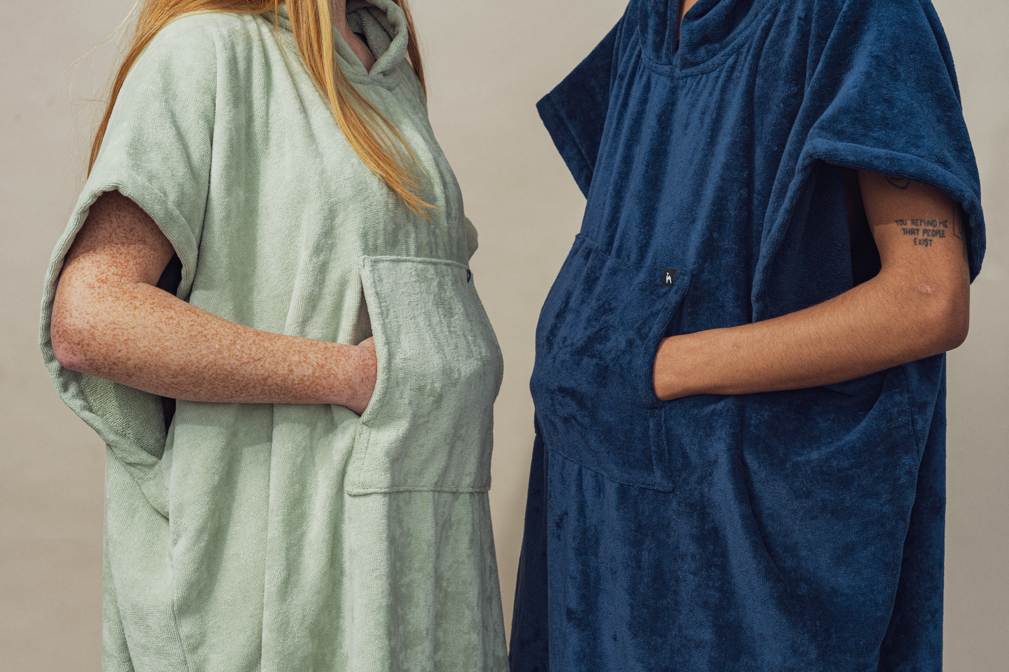 Two people stand side by side, wearing Futah's Ericeira Terry Poncho—one in light green, one in dark blue. The made-in-Portugal cotton robes feature large front pockets; they face each other against a neutral background.