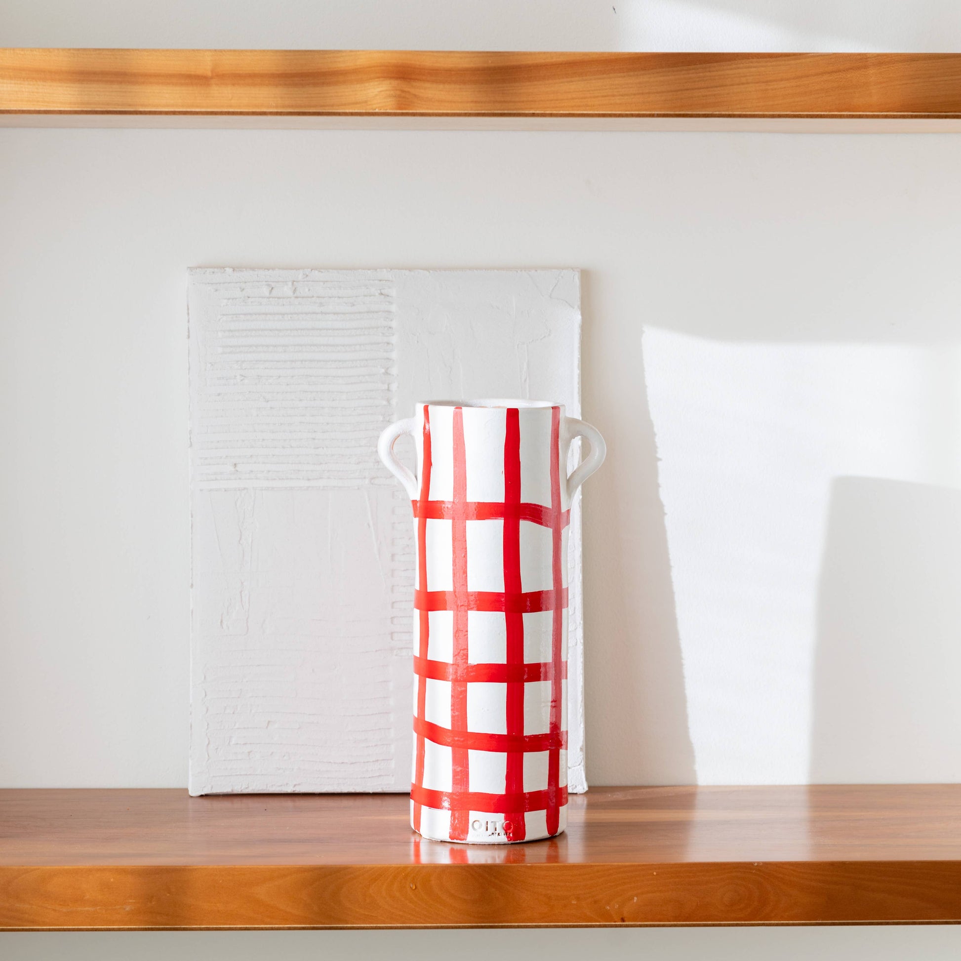 The Udine vase by OITO Atelier—a tall white handmade ceramic piece with red grid lines and two small handles—sits on a wooden shelf before a textured white canvas, sunlight casting shadows for an artisanal decor accent.