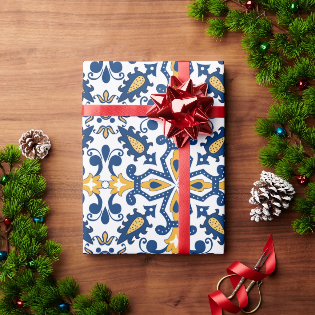 A gift wrapped in blue, yellow, and white paper with a red bow rests on a wooden table amidst pine branches, pinecones, ornaments, and the Iberica - Pretty things from Portugal Iberian sampler kit - Provenzal for a festive holiday scene.