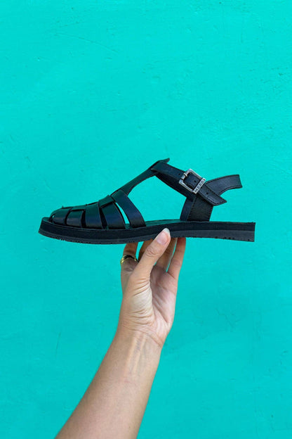 A hand displays the Black Glory sandals by Iberica - Pretty things from Portugal, featuring a buckle design against a vivid turquoise background, highlighting their comfort and versatility.
