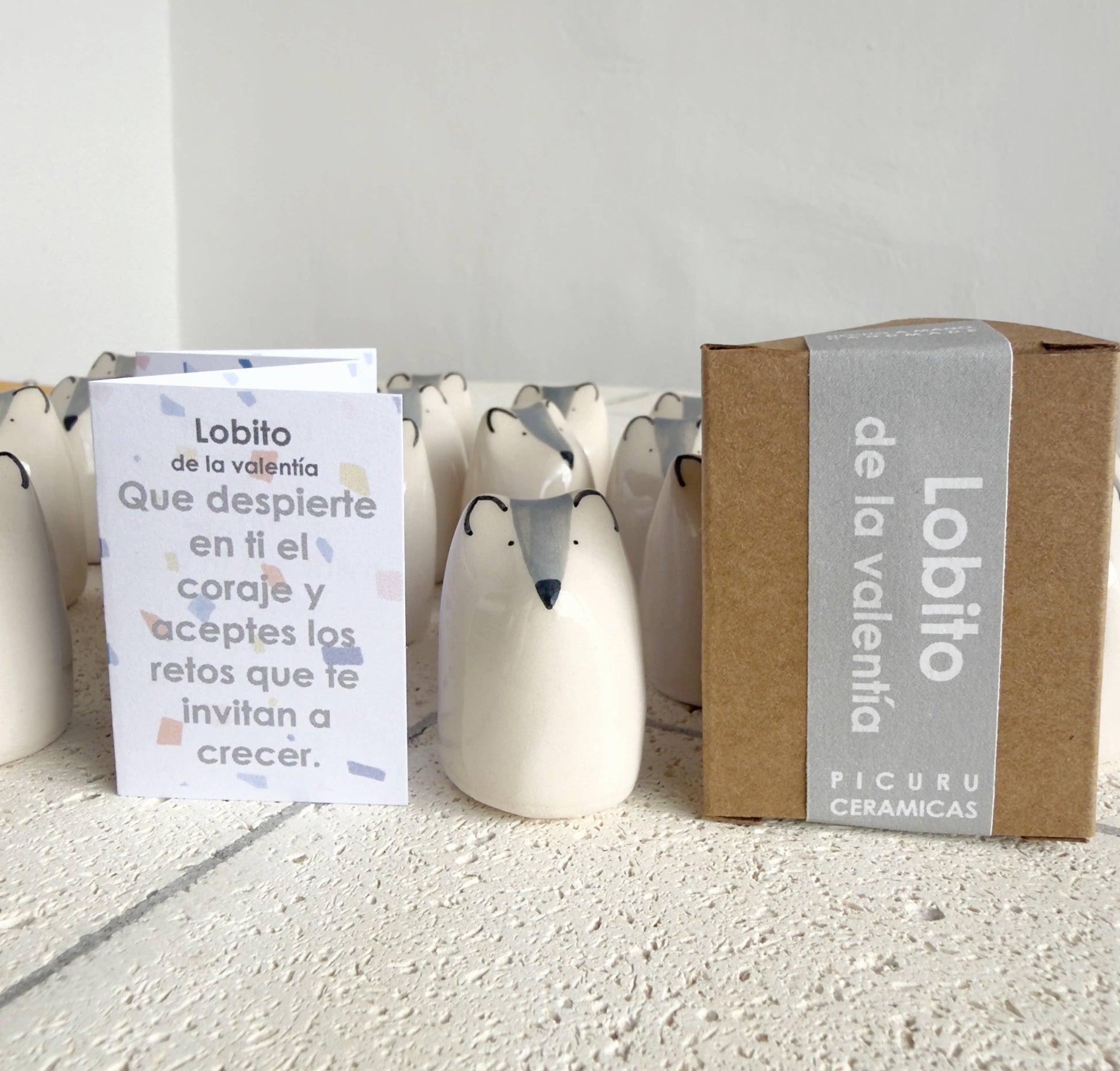 The Little Wolf Amulet of Courage by Picuru ceramicas, a handmade ceramic symbol of strength and growth, stands on a white surface beside its “Lobito de la valentía” box and an inspirational Spanish card; identical figurines are in the background.