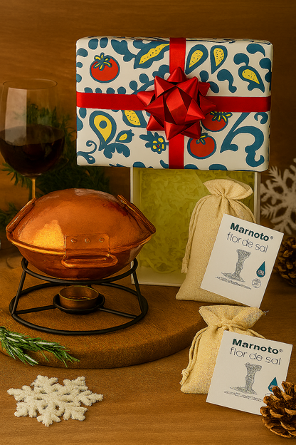 The Cataplana starter kit by Iberica - Pretty things from Portugal includes a festive wrapped present, red wine glass, copper cataplana on a stand, two Marnoto flor de sal bags, pinecones, greenery, and a crocheted snowflake—ideal for Portuguese cooking.