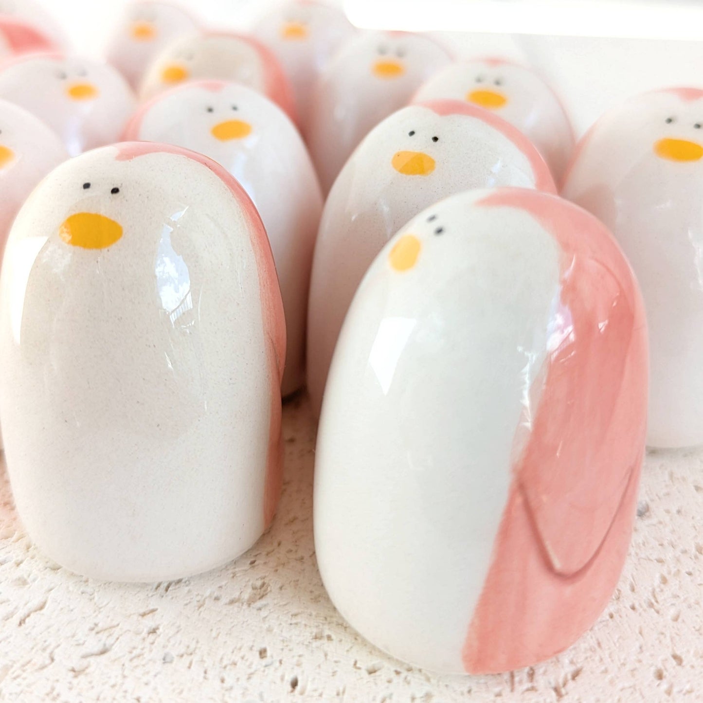 A cluster of Picuru ceramicas’ Penguin Amulets of Presence—handmade ceramic figurines with white, pink, and yellow details—rest on a textured surface, each symbolizing balance and resilience.