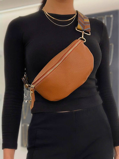 A person wears a fitted black long-sleeve shirt and gold necklaces, styled with the Cassette Barcelona Leather Fanny Pack Crossbody Bag featuring a wide patterned strap. The background is softly blurred.