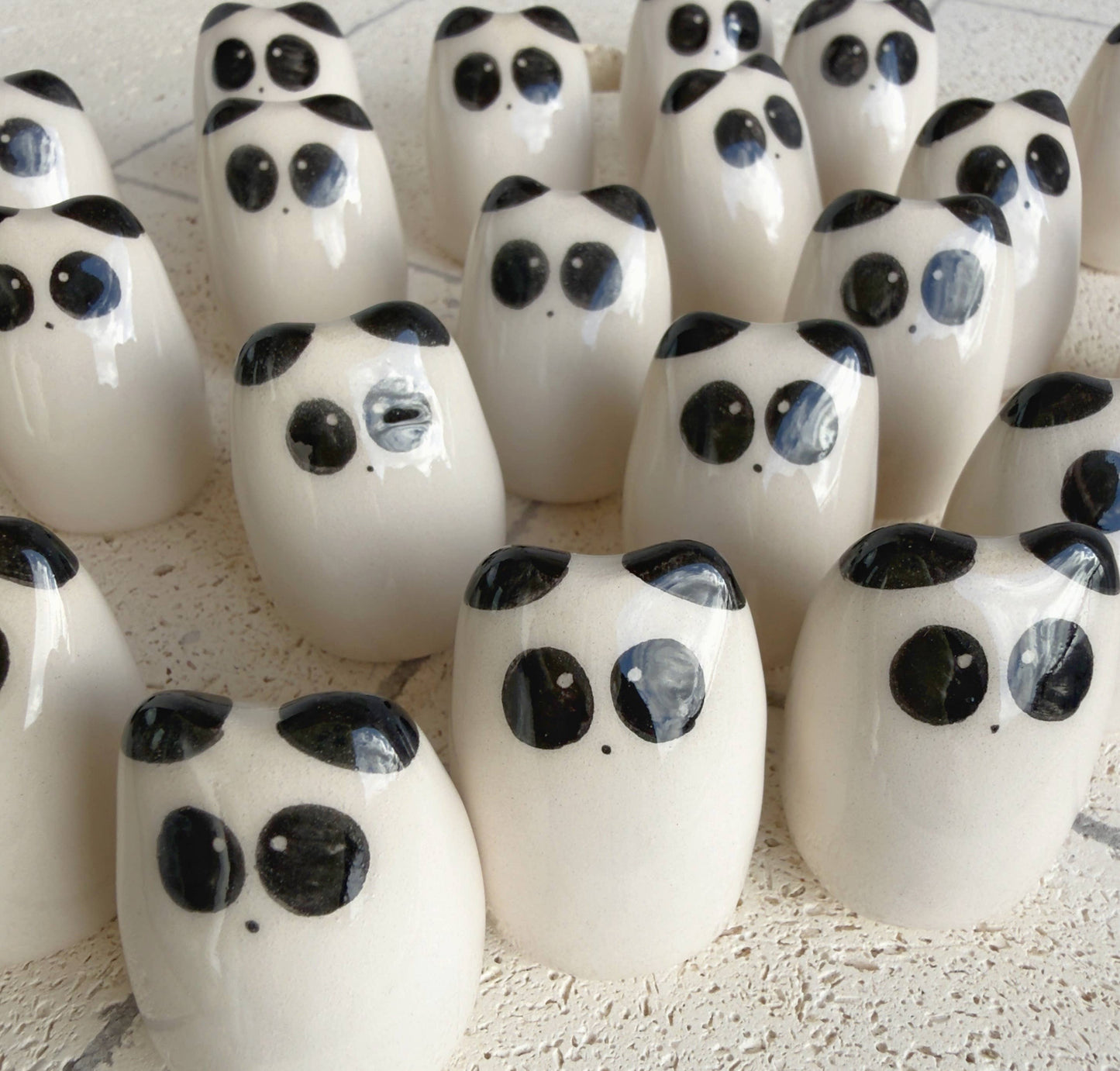 A cluster of small, glossy white figurines with black eyes and ears, resembling minimalist pandas by Picuru ceramicas, forms the Panda Amulet of Inner Peace – Handmade Lucky Charm Gift, arranged closely on a textured light surface.