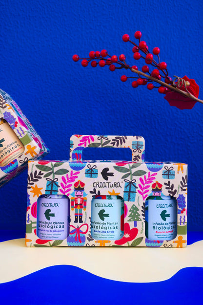 The Christmas Tea Box | Criatura X Teresa Rego - Special Edition features three bottles of Criatura organic herbal teas in a festive, floral box with a Nutcracker design—an ideal holiday gift. Red berries and bright decor accent the blue wall backdrop.